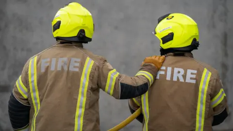 PA Media The backs of two firefighters wearing brown fire jackets and wearing yellow helmets. One is holding a yellow hose under their left arm while the other has their hand on the other's shoulder.