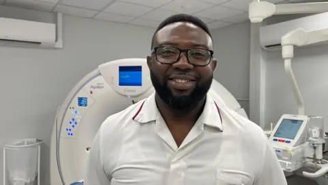 Bempah photographed looking into the camera and smiling. He is wearing a white collared shirt with a dark red trim around the collar, and dark-framed glasses. He has short, dark curly hair and a short beard. He is pictured inside an MRI scanning room, with the machine - a large, white, doughnut-shaped structure - behind him. The machine has an array of blue lights on it, and a screen at the apex. The room has dark grey walls and a white ceiling. 