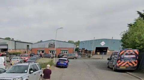 Coder Road at Ludlow Business Park with warehouse buildings and parked cars. A road leads up to several large warehouses with brick lower sections and corrugated steel upper portions.