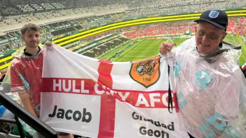 Jacob Allmendinger Two men are holding a flag in a large football stadium, they have rain coats on over their England football shirts. On the left, a young man with brown hair and a closely cropped beard wearing a red shirt. On the right, an older man wearing a blue baseball cap with England "Three Lions" logo and a white shirt. The flag is a red and white flag of St George, with the words "Hull City AFC written across the centre, the names "Jacob" and "Grandad Geoff" in the lower quadrants and a Hull City tiger badge in the upper right quadrant. 