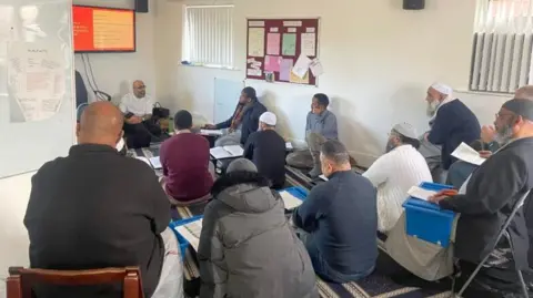 A small group of men sit in a room with white walls. There is a noticeboard on one wall and windows covered by blinds.  There is a television screen at one end, under which a man in a white top is sitting to deliver a training course.  Some of the men are wearing skull caps.