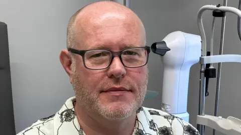 A man wearing glasses and a white shirt with black flowers on it. He is in a room at an optician surgery.