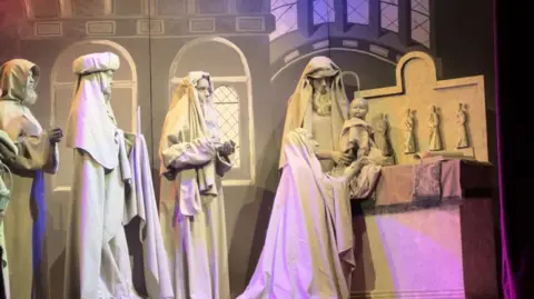 Five female students on stage recreating a nativity scene, head to toe in grey costumes, with grey paint on their faces, hair and hands