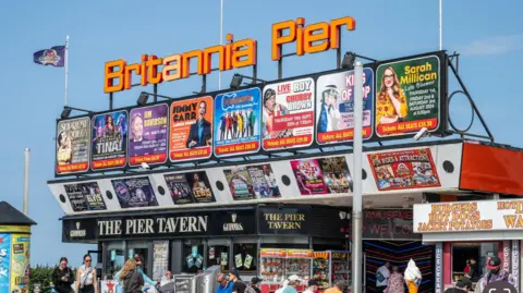 Andrew Turner/BBC View of Britannia Pier from the front on the right. A orange sign is above the entrance that says Britannia Pier, and multiple posters that show who is set to be at the pier.