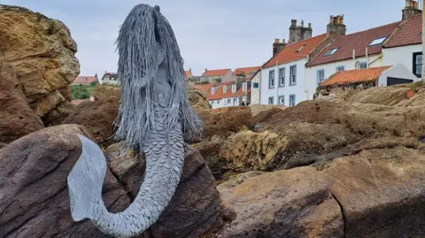 George Carson A grey mermaid sculpture, sitting on rocks on a beach, with houses visible in the distance. 