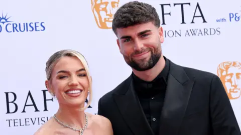 Molly Smith and Tom Clare pose together. Molly is wearing a silver necklace and off-the-shoulder dress and Tom is wearing a black suit over a black shirt. 