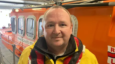 A man, wearing a yellow jacket and red lifejacket, standing in front of an orange RNLI boat. 
