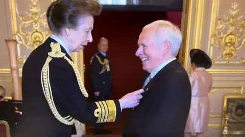 The Princess Royal wearing ceremonial uniform pinning the MBE on Nick Owen's suit. He is wearing a dark suit and silver tie and is smiling. 