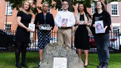 Four women and one man stand alongside a large rock showing a memorial plaque. They are all holding photos of relatives killed in road crashes.