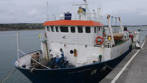 The Gry Maritha has a blue hull and a white wheelhouse . Rust stains are visible all over the vessel