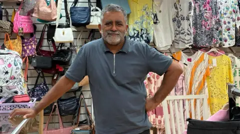 LDRS A man with short grey hair and a beard is wearing a grey polo shirt. He is smiling while standing in his stall. Surrounding him are various items of clothing and handbags.