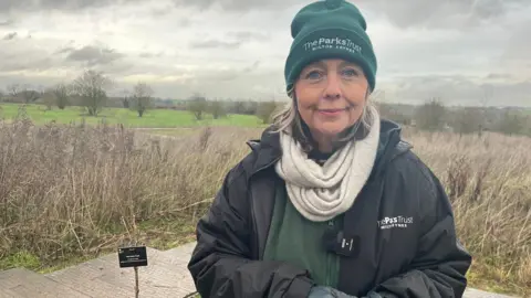 Nicola Haseler/BBC Victoria Miles smiles at the camera, standing next to the sapling which is barely visible against the brown grass and brown footpath behind it. She wears a Parks Trust black anorak over a green rangers fleece, and a matching green Parks Trust beanie hat. Behind her is grassland and short bare trees dotted around. The sky is cloudy and leaden.