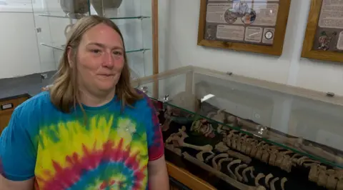 A close-up of a woman, Dr Eleanor Blakelock, wearing a blue, yellow and red top. She has shoulder-length light brown hair. She is standing in front of a case full of bones.