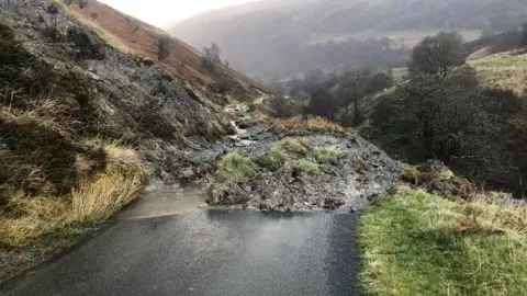 Rural minor road blocked by large mound of mud and stones which have slipped down from bank to the left