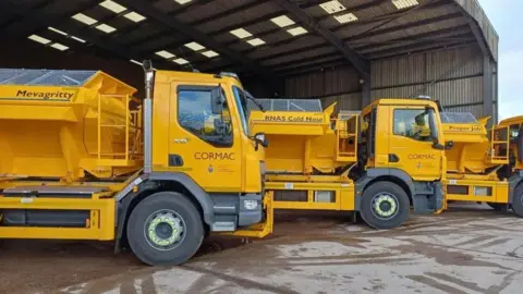 Three bright yellow gritting lorries lined up outside an open-fronted warehouse at a depot.