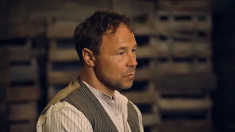 Actor Stephen Graham wearing 1930s clothing in a promotional shoot for Peaky Blinders. He has short, dark hair and stubble on his face.