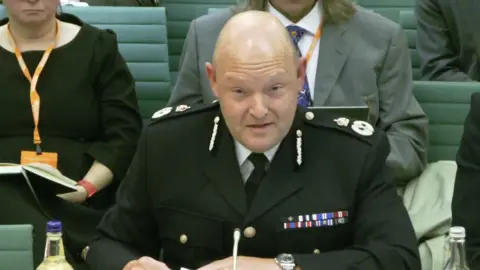 Former Chief Constable Craig Guildford sits at a desk while wearing a black police uniform with a medal ribbon on it. Several people are sitting on green chairs behind him.