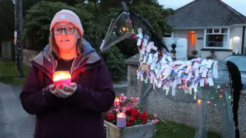 A woman standing outdoors in the evening, holding a lit candle in both hands. She is wearing a light-coloured beanie, glasses, and a purple coat. Behind her is a wireframe sculpture of a horse, adorned with colourful notes, woollen tags and small string lights.