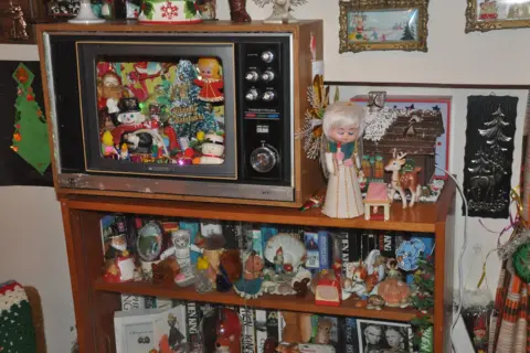 A vintage wooden television cabinet decorated for Christmas, with the TV screen filled with colourful Christmas figurines and lights, and shelves below crowded with books and assorted collectibles, including dolls, animals, shells, and small ornaments.