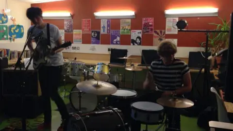 Josh Law/Ben Sadler The pair playing a guitar and drums inside a dark room with an orange wall.