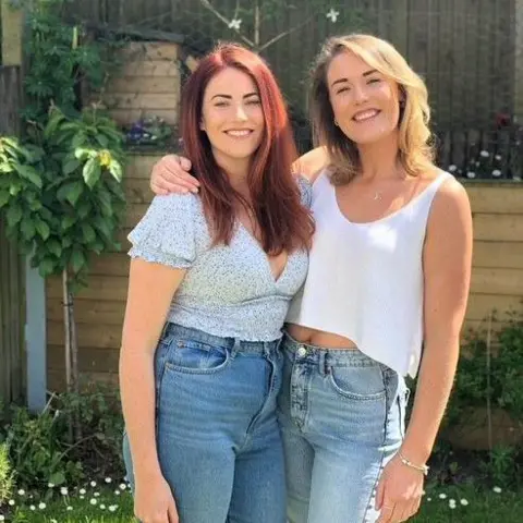 Leanne and Sarah/BBC Leanne (left) and Sarah (right) smile as they stand in a sunny garden. Leanne has long red hair and Sarah has light shoulder length hair. 