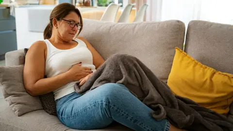 Getty Images A woman sits on the sofa at home with a blanket on top of her and yellow cushion to her side, clutching her stomach.