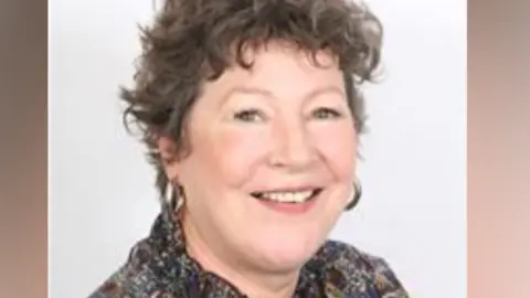 Sue Riley - a middle aged white woman with short, brown, curly hair. She is posing for a staged photo in front of an all-white background and is wearing a predominantly brown multi-coloured shirt. She has large hooped earrings on and is smiling
