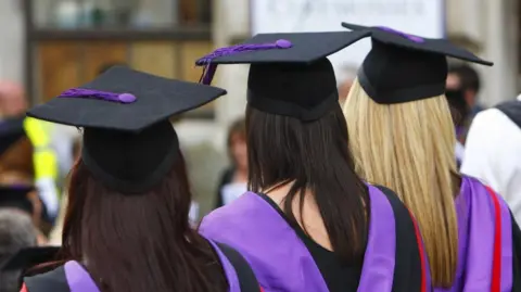 Students wearing a black graduation cap. They have long hair and a black gown with purple detailing.