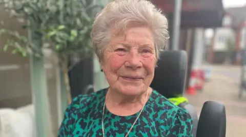Julia Gregory/BBC Norah Holder is sitting in a chair wearing a green and black patterend top and a cross on a chain around her neck. She has short white hair and is wearing ear studs and smilling at the camera, turned slightly sideways. There is foliage behind her.
