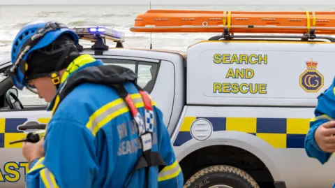 HM Coastguard Search and Rescue members stand next to an emergency vehicle. They wear blue and yellow uniform and a helmet.