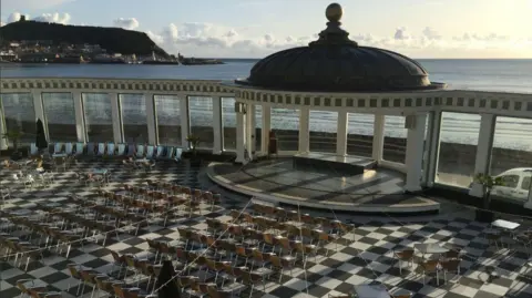 BBC/Julia Lewis Seating laid out on a black and white checked floor space inside Scarborough Spa.. The headland, sea and beach are in the background.
