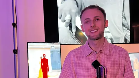 Aleks Tamasauskas at Chet Lo's London Fashion Week show. Mr Tamasauskas wears a pink and brown checked shirt and he holds a grey cane with a black top. He stands in front of three screens showing images from London Fashion Week