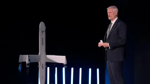 Andrew Matthews/PA Wire Alistair Carn standing on a stage giving a speech in a suit and tie. He has a model of a drone next to him