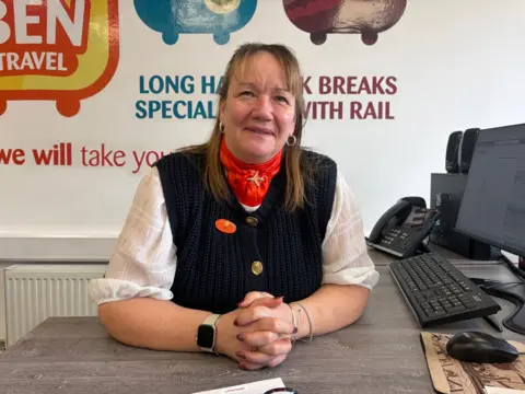 Sue sits behind an office desk. She is wearing a white shirt with her sleeves rolled up and a navy gillet-styled jumper. She has a red neck scarf. 