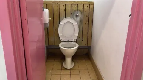 Toilet cubicle and the door and frame is pink. The floor is a bit dirty