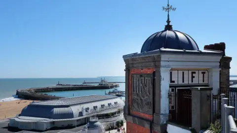 An Edwardian lift overlooking a seafront with a domed roof and tiled sides. Beyond it are the sea, blue sky, a curved promenade and a beach.