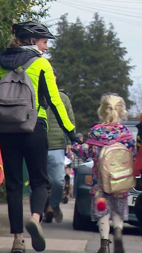 A parent wearing cycling safety gear holding the hand of a child whilst walking to school