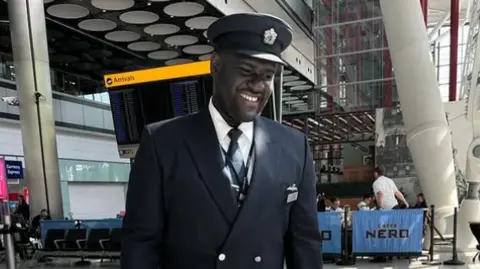 Yeboah in his British Airways uniform which includes a navy suit and hat. He is smiling and looking off-camera.