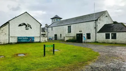 David Nixon Two separate white buildings, one featuring a large bird artwork, with an access track and some grass in the foreground