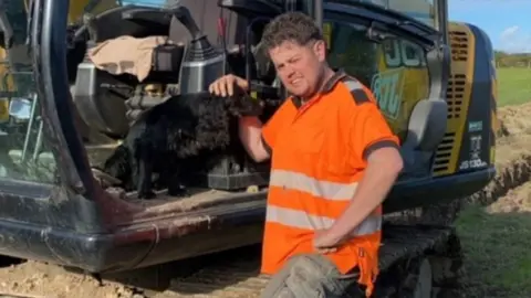 Man in an orange hi-vis stroking his dog who is in a JCB