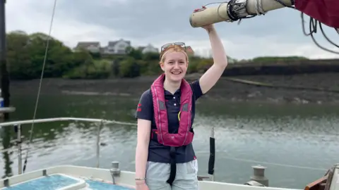 Young woman with a pink life jacket on holds onto part of the rigging of the boat smiling