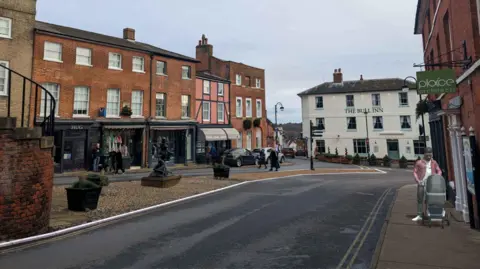 Suffolk County Council An artist's impression of Seckford Street in Woodbridge. Businesses and houses line each side of the road. A pub is in the distance. A man walks a pram down the pavement.