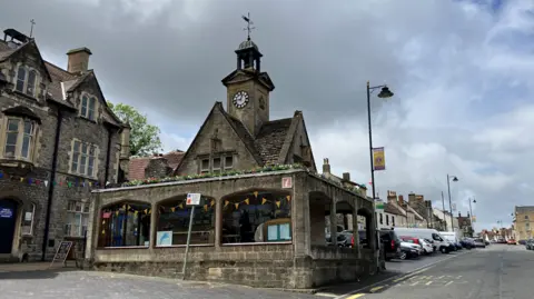 A stone clock tower, with a weather vane on top, stands at the edge of a high street. There are flower boxes on the upper level. Its lower section features a partially glazed arcade. Rows of parked cars can be seen in the distance and to the left is a three storey brick building. 