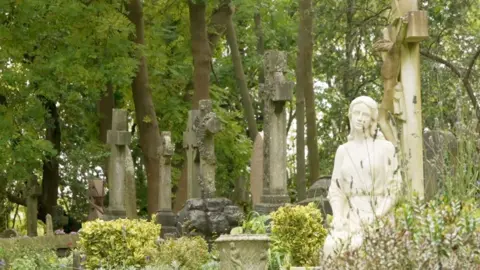 Religious gravestones, with crosses on the headstone surrounded by tall trees. There is a white statue of an angel in the foreground. 