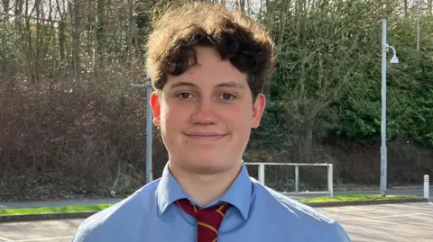 Grace Wood/BBC A man with short curly brown hair wearing a blue shirt and red and yellow striped tie stands on a petrol forecourt