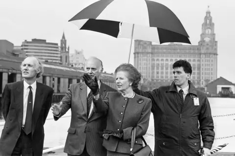 Getty Images Black and white photo of Margaret Thatcher in front of the Liver Building in Liverpool in 1989, pointing with one hand, next to two men in suits and another man holding an umbrella above her.