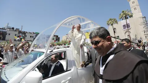 Getty Images The pope, waving to people, stands in the white popemobile, smiling