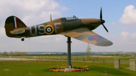 Geograph/Christine Matthews A model Hurricane Hawker held aloft by a large grey stand. The plane is decorated with green and brown camouflage and stands in front of fields at the airfield. 