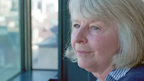 A women with straight fair hair and blue eyes looks out of a window. There are buildings, out of focus, in the background. 