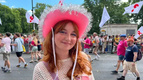 A medium close-up of a smiling attendee named Magda Szewczak, who has red hair and glitter eye makeup, wearing a bright pink cowboy hat trimmed with silver tinsel at the parade.